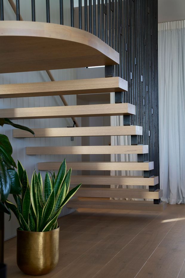 Interior view of a contemporary floating staircase with broad light timber treads and a curved upper landing. Slim dark vertical metal balusters form a striking screen beside the stairs, with pale vertical wall panelling, full-height sheer curtains, and a large potted snake plant in a brass-toned planter. The stair void and open risers create an airy, sculptural composition.