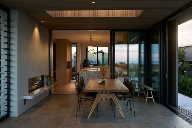 A refined open-plan dining and living area with a long solid timber dining table, black metal dining chairs, and a sculptural timber bench. The room features a linear fireplace set into a pale textured wall, a warm timber slatted ceiling with a large recessed skylight or clerestory opening, and extensive black-framed glazing and sliding doors. A compact kitchen is visible beyond the dining area, with a glimpse of landscaped exterior space through the glazing. The palette is warm and earthy, combining timber, stone-toned flooring, dark metal, and soft neutral finishes.