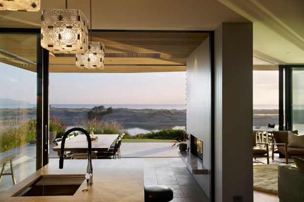 Open-plan contemporary living space with a kitchen island in the foreground, a dining area beyond, and a lounge visible to the right. Full-height glazing frames a coastal landscape, while warm timber ceiling lining, dark-framed openings, a tiled floor, and sculptural pendant lights create a refined indoor-outdoor connection.