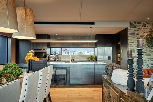 A sophisticated open-plan kitchen and dining/living area. The kitchen features dark charcoal built-in cabinetry, a pale stone-look benchtop and splashback, stainless-steel appliances, and a large island with stools. Warm timber flooring, oversized woven pendant lights, leather and upholstered seating, and a partially exposed rough-stone wall create a refined, textural interior. Daylight enters through large windows, with neighbouring rooftops visible beyond.