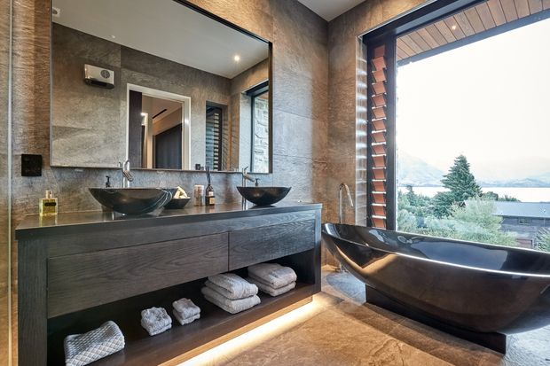 A luxurious contemporary bathroom with a double vanity, two black vessel basins, a dark timber floating vanity, large wall mirror, and freestanding black bathtub. The room features warm stone-textured wall and floor finishes, recessed lighting, and a large louvred window offering views toward a lake or waterfront landscape.