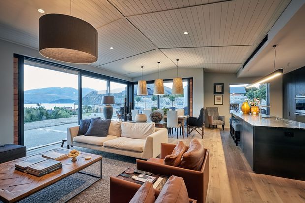 A spacious open-plan contemporary living, dining and kitchen interior with extensive glazing overlooking a mountainous waterfront landscape. The room features a light timber-lined ceiling, wide timber flooring, a neutral sectional sofa, leather lounge chairs, a timber coffee table, a dining area with woven pendant lights, and a dark kitchen island with integrated cabinetry. The interior has a warm, refined feel and strong indoor-outdoor connection.