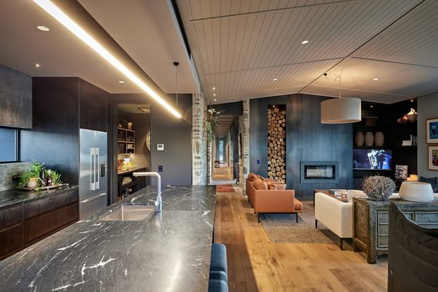Open-plan contemporary kitchen and living area with a large dark stone island and integrated sink, dark timber cabinetry, stainless-steel refrigerator, timber flooring, and a textured timber-lined ceiling. The living area includes a fireplace set into dark wall joinery, tan and cream seating, a large pendant light, shelving, and a decorative stacked-log feature beside the hallway.