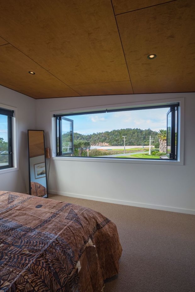 A bright bedroom with a partially visible bed, patterned brown bedding, carpeted floor, pale walls, and a warm timber-panelled ceiling with recessed downlights. Large black-framed awning windows are open to a green, semi-rural outlook, and a full-length mirror leans against the wall beside the bed.