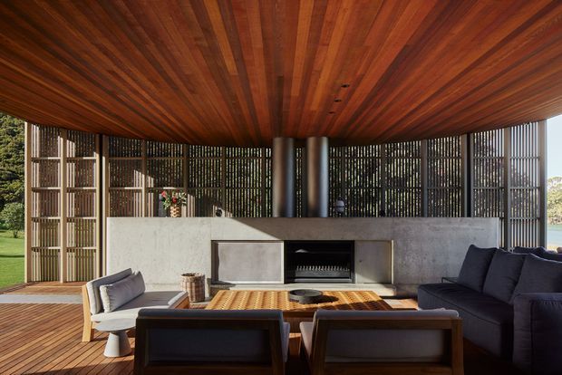 A sophisticated covered outdoor lounge with a warm timber-lined ceiling and deck, framed by vertical slatted screens overlooking landscaped grounds and water. The space features a concrete fireplace wall with a built-in firebox, dark contemporary sofas, upholstered lounge chairs, and a low timber coffee table. The open sides create a strong indoor-outdoor connection with abundant natural light.