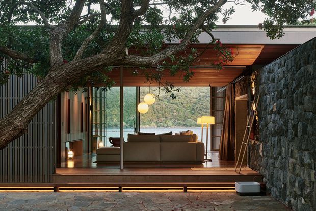 A refined open-sided living pavilion framed by a large tree and a dark natural-stone wall. The room features a timber-lined ceiling, full-height glazing, vertical timber screening, warm concealed lighting, and a neutral sofa facing a wooded waterside landscape. A stone-paved terrace or courtyard occupies the foreground.