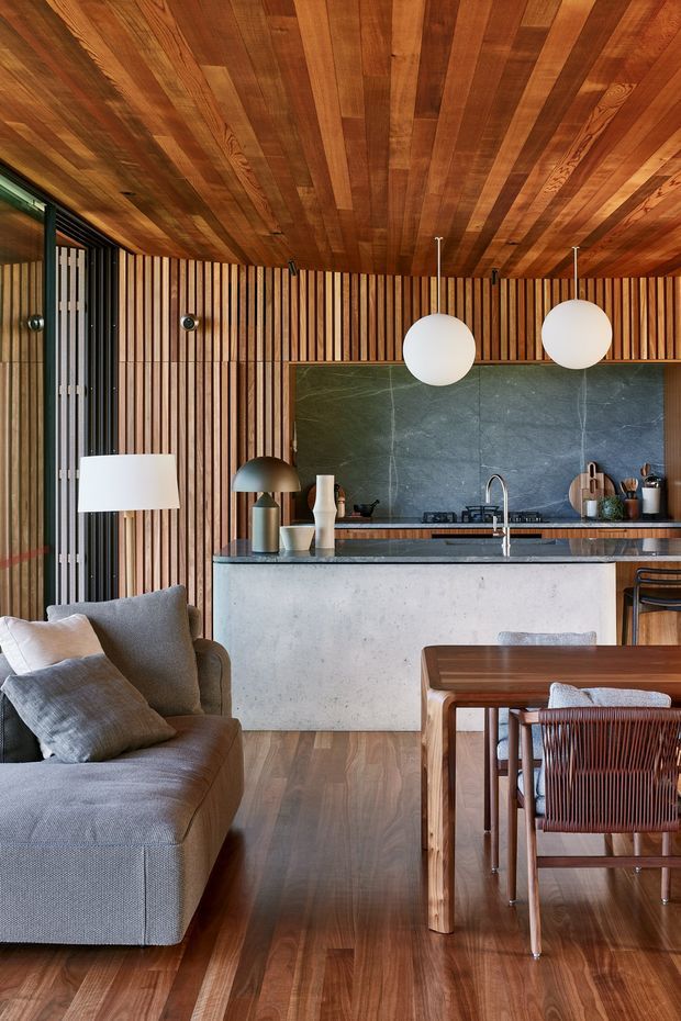 Warm open-plan interior combining a living room, dining area, and compact kitchen. The space features extensive vertical timber battens on the walls, a timber slat ceiling, timber flooring, a dark stone splashback, and a pale concrete island. A grey upholstered sofa occupies the foreground, with a timber dining table and woven dining chairs to the right. White spherical pendant lights and a floor lamp provide contemporary lighting.