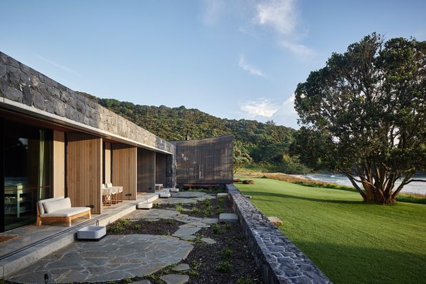 A low, modern stone-clad residence set beside a landscaped lawn and waterfront. The foreground shows a broad irregular stone-paved terrace, timber-framed floor-to-ceiling glazing, vertical timber screening, and restrained outdoor seating. A low stone retaining wall borders the lawn, with dense wooded hills and water beyond. The composition has a strong connection between the house, garden, and natural setting.