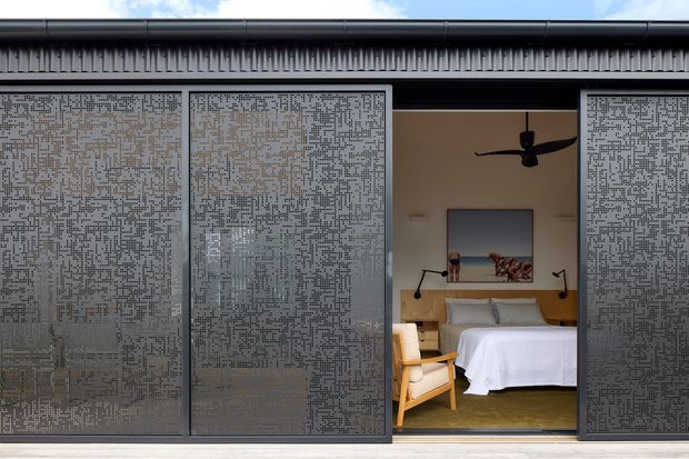A contemporary single-storey facade with large patterned perforated metal sliding screens partially enclosing an open bedroom. The bedroom contains a neatly made bed, warm timber-framed armchair, wall-mounted reading lights, artwork, and a dark ceiling fan. The composition emphasises privacy screens, indoor-outdoor connection, and a restrained palette of charcoal grey, white, timber, and muted yellow.