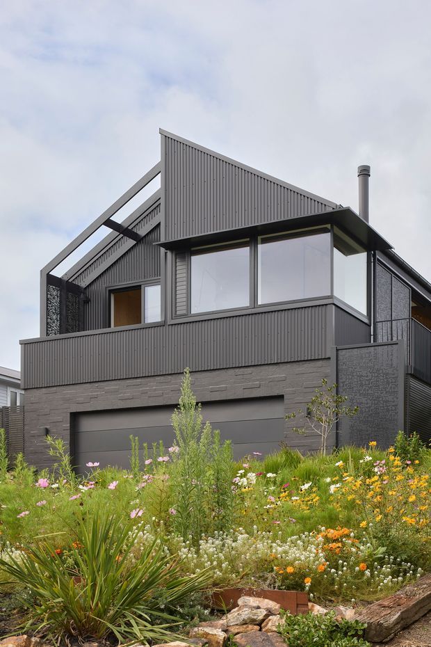 Exterior view of a contemporary multi-level house with dark grey vertically profiled cladding, a steep asymmetrical gabled roof form, large upper-level glazing, a projecting balcony or terrace, and an integrated double garage. The lower facade uses grey masonry, while a densely planted garden with wildflowers and rock edging fills the foreground.