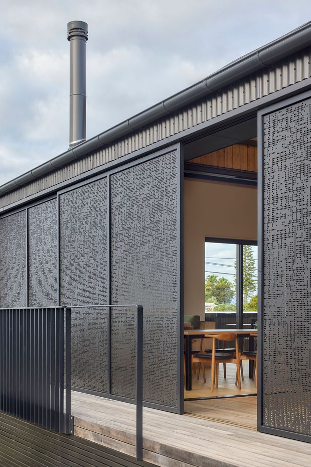 Exterior view of a contemporary low-pitched-roof building with dark grey perforated sliding screens, corrugated metal wall cladding, charcoal gutters and a cylindrical flue. A timber deck and dark metal balustrade lead to an open doorway, revealing a warm timber-lined dining area with a table and chairs inside.