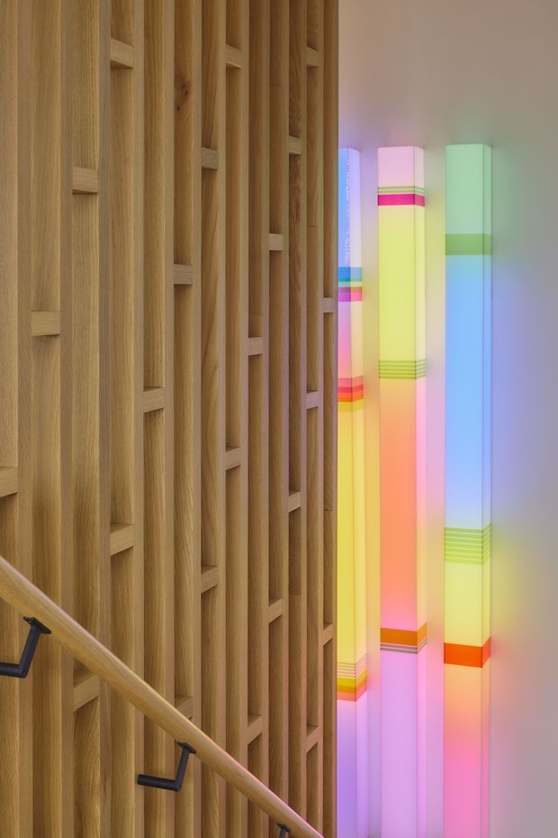 Interior architectural detail showing a timber-clad stairway or landing. The wall is composed of vertical natural timber battens with recessed rectangular detailing, accompanied by a timber handrail with dark metal brackets. Beside it are tall translucent illuminated columns with vivid multicolour bands, creating a contemporary sculptural lighting feature.