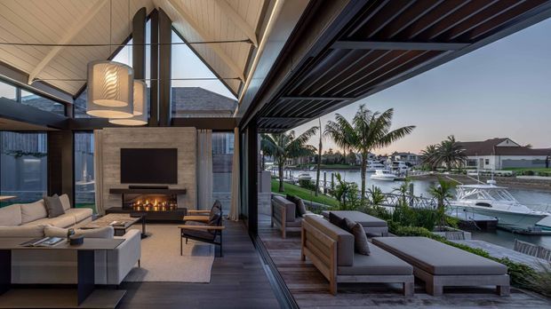 A sophisticated open-plan living room opening onto a covered outdoor lounge terrace, with expansive retractable glazing framing a waterfront canal and boats. The interior features a suspended fireplace set into a textured stone or concrete feature wall, pale upholstered seating, dark timber flooring, sculptural pendant lights, and a high pitched ceiling with exposed structural beams. The outdoor terrace continues the dark timber palette with built-in-style lounge seating, a timber slatted overhead canopy, and lush palms beyond.