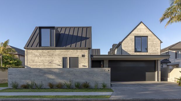 Contemporary two-storey residential facade viewed from the street, with pale horizontal masonry or concrete-block walls, a dark metal-clad upper volume, prominent steep gable roof, large dark-framed windows, and an integrated double garage. A low rendered boundary wall, narrow landscaped planting strip, driveway, and palm trees frame the front elevation.