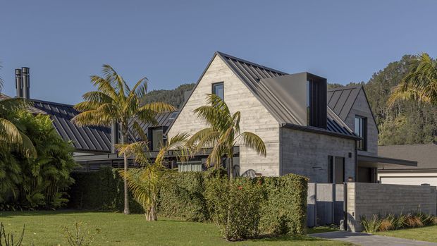 Exterior view of a contemporary multi-level residential home with steep gabled roof forms, dark standing-seam metal roofing, pale horizontal masonry or stone-look wall cladding, and dark-framed windows. The property is landscaped with palms, manicured hedges, lawn, and a paved driveway, with neighbouring structures and a wooded hillside in the background.