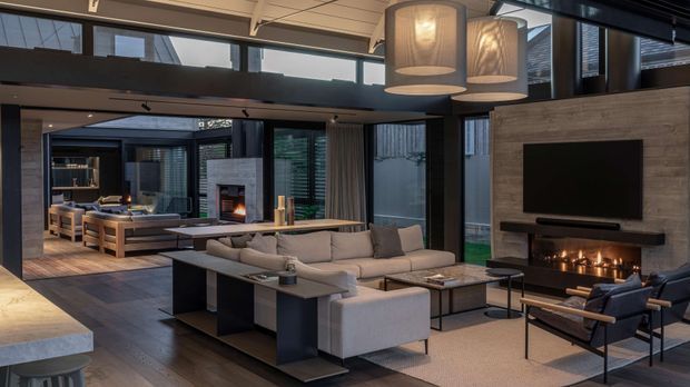 A spacious, sophisticated open-plan living room with a double-height ceiling and mezzanine-like upper glazing. The room features a large sectional sofa, dark-framed floor-to-ceiling windows, a linear fireplace beneath a wall-mounted television, contemporary pendant lights, timber flooring, and a secondary lounge and fireplace visible beyond. The palette is warm and restrained, combining timber, charcoal, grey, and cream finishes.