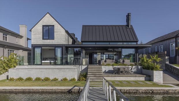 Exterior view of a contemporary multi-level residence beside a narrow water feature or canal. The home combines a steep gabled upper volume clad in pale material with a dark standing-seam metal roof over a lower glazed living area. Broad sliding glass doors open toward a stepped terrace with concrete retaining walls, outdoor seating, planted garden beds, and a small footbridge in the foreground. A neighbouring residential building is visible to either side under a clear blue sky.