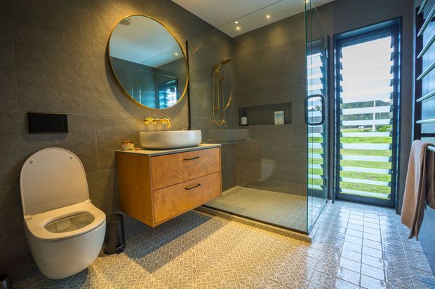 A contemporary bathroom with a wall-mounted toilet, floating timber vanity with a white vessel basin, round brass-framed mirror, wall-mounted brass tapware, and a frameless glass walk-in shower. Dark textured wall tiles contrast with patterned floor tiles, while louvred glazing brings in bright natural light and views to greenery.