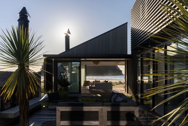 A modern dark-clad coastal house viewed from an outdoor deck or terrace, framed by spiky architectural planting. The angular gabled roof and strong vertical cladding create a contemporary silhouette, while large openings lead to a living area and reveal a sandy beach and ocean beyond. Outdoor lounge seating and built-in planters occupy the foreground.