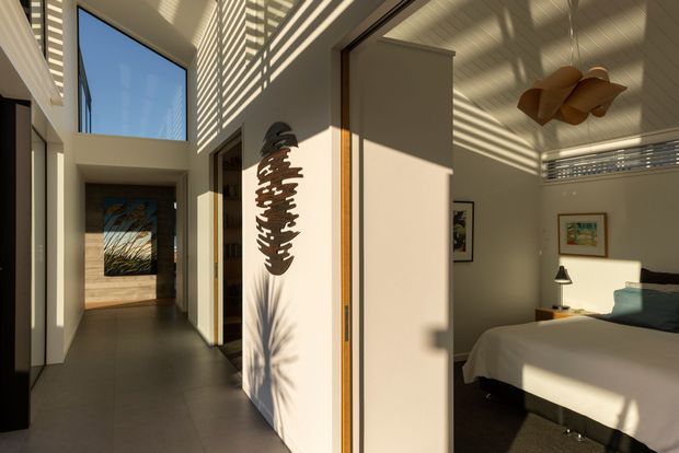 Sunlit contemporary bedroom viewed from an adjoining hallway, with a high ceiling and clerestory-style glazing casting strong horizontal shadows across pale walls. The room contains a neatly made bed, bedside table and lamp, framed artwork, and a sculptural timber pendant. A large internal pivot or sliding door separates the bedroom from the corridor, while a double-height opening and upper window bring in abundant natural light.
