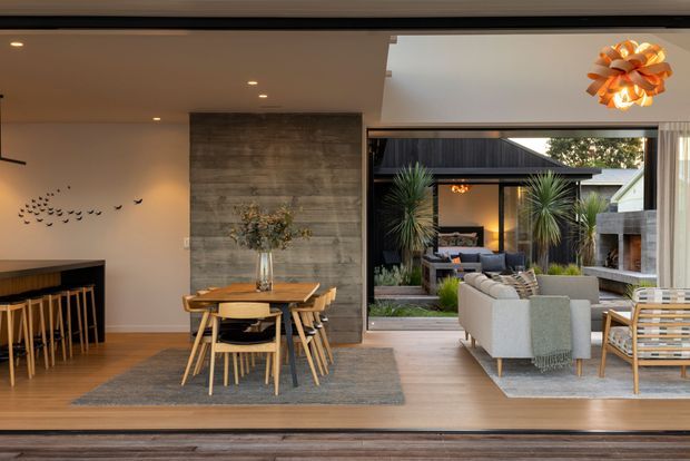 A refined open-plan interior combining a dining area, kitchen or bar counter, and living room, opening broadly to a landscaped outdoor courtyard. The room has warm timber flooring, a textured concrete feature wall, pale plaster walls, timber dining furniture, a large neutral rug, and contemporary upholstered seating. Full-width glazed sliding doors create a strong connection to the outdoor entertaining area, which includes planting, a covered structure, and an outdoor fireplace. Recessed ceiling lights and a sculptural pendant provide layered lighting.