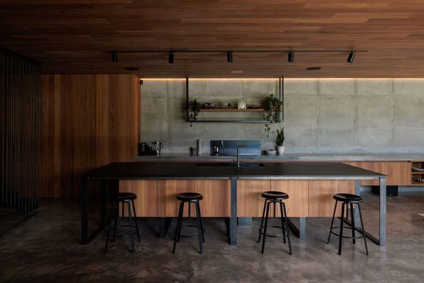 A dark, refined contemporary kitchen with a large central island and four bar stools. The room features extensive warm timber panelling and a timber ceiling, a dark benchtop, concrete-look wall and floor finishes, black metal detailing, integrated track lighting, open shelving, and understated decor.