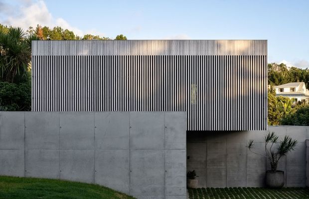 Exterior view of a modern, rectilinear house concealed behind a tall vertically slatted facade. The upper volume has closely spaced dark vertical battens with a weathered metallic or timber-like appearance, while the lower boundary and retaining walls are smooth exposed concrete. A recessed entry or undercroft sits behind the wall, with patterned grass-and-paver landscaping and sculptural planting. The composition is minimalist and strongly geometric, with a flat roof and partially elevated upper level.