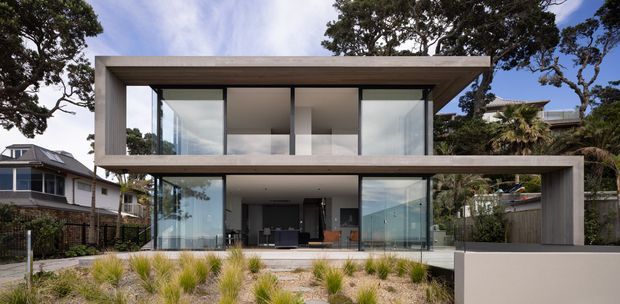 A contemporary two-storey residence with a strong rectangular composition, flat roof, deep projecting concrete or rendered frames, extensive floor-to-ceiling glazing, and dark window framing. The house opens toward a landscaped front garden with ornamental grasses and is surrounded by established trees and neighbouring homes.