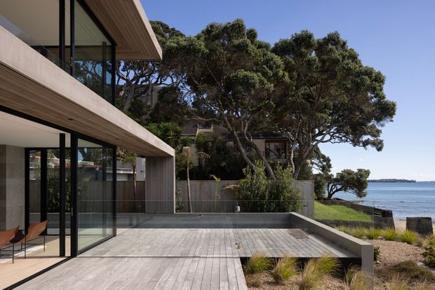 A contemporary multi-level coastal residence with broad cantilevered timber-lined roof and balcony overhangs, expansive black-framed glazing, and a large timber deck stepping toward a landscaped waterfront edge. Sculptural coastal trees, grasses, a calm bay, and distant shoreline are visible beyond the terrace.
