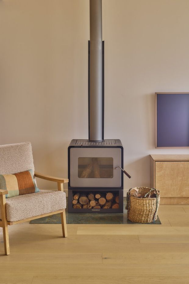 A warm, softly lit living room centered on a freestanding wood-burning stove with a tall flue and integrated log storage. The room has pale timber flooring, beige walls, a textured upholstered armchair with a multicoloured cushion, a low timber media cabinet, and a woven basket. The composition is calm and inviting, with a simple contemporary cabin-like feel.
