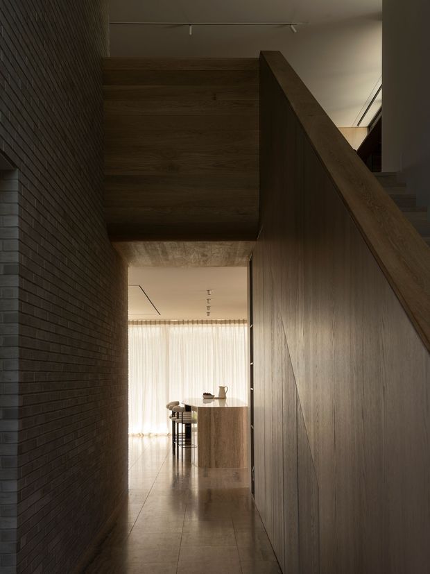 A narrow, moody residential hallway viewed toward a bright dining and kitchen area. The corridor is framed by a dark exposed brick wall on the left and extensive warm timber wall panelling and stair balustrade on the right. A timber kitchen island with stools is visible in the background, with sheer curtains providing soft daylight. The space has polished hard flooring, subdued linear lighting, and a refined contemporary architectural character.