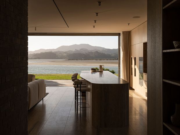 A warm, refined open-plan kitchen and living area with a large dark stone island and bar seating in the foreground. Floor-to-ceiling sliding glazing opens directly toward a waterfront landscape, with mountains visible beyond the water. The interior features timber-toned built-in cabinetry, dark brick wall cladding, a tiled floor, recessed ceiling lighting, and a neutral palette.