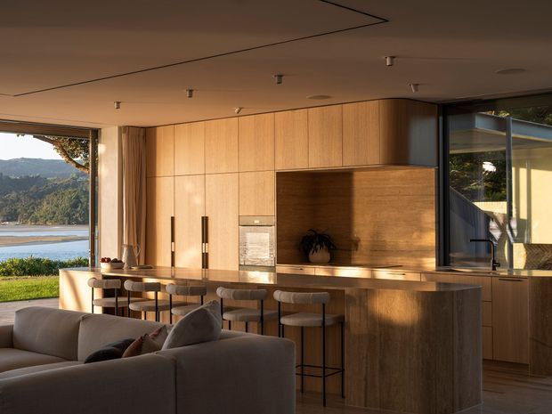 A warm, open-plan contemporary kitchen and living area with extensive light timber cabinetry, a long island bench with integrated seating, and a recessed timber feature niche. Large glazed openings connect the room to a lush waterfront landscape and provide abundant natural light. A neutral sofa occupies the foreground, while minimal ceiling details and concealed lighting create a refined, streamlined interior.