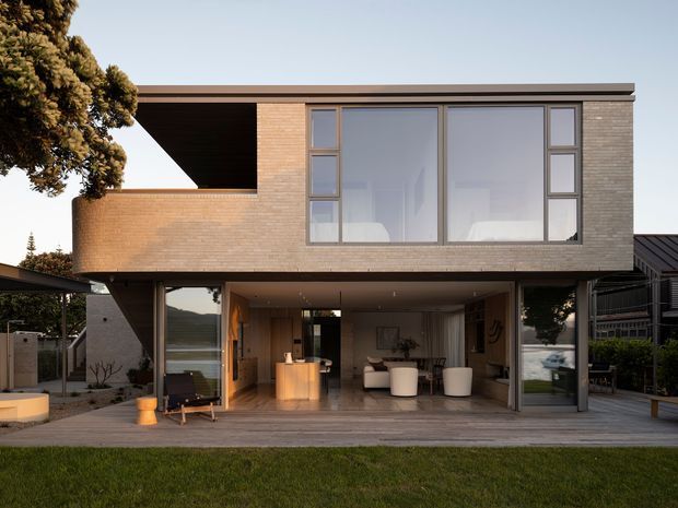 A contemporary two-storey residence viewed from the rear, with a cantilevered upper level, flat roof, warm brick walls, extensive floor-to-ceiling glazing, and an open ground-floor living area connecting to a timber deck and lawn. The sheltered outdoor living zone contains lounge and dining furniture, while the interior kitchen and living room are visible through the glazing.