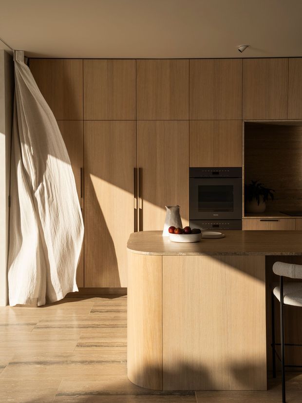 Warm, minimalist kitchen interior with extensive floor-to-ceiling light timber cabinetry, integrated dark oven, rounded kitchen island with timber cabinetry and benchtop, and a partially visible bar stool. Strong low-angle sunlight casts dramatic shadows across the cabinetry and timber floor. A sheer curtain hangs beside the cabinetry, and a recessed open shelf contains a plant.