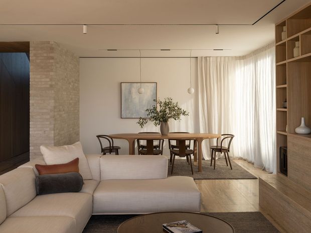 A warm contemporary open-plan living and dining area. A large cream sectional sofa faces a dark built-in media wall and timber shelving, while a timber dining table with curved wooden chairs sits behind it. Pale textured masonry forms a feature wall, with full-height sheer curtains, a neutral rug, pendant lights, track lighting, and a muted artwork completing the calm, refined interior.