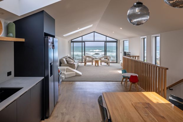 A bright open-plan coastal home interior combining a modern kitchen, dining area and living room. The foreground features a timber dining table and chairs, while the living area beyond has a light sectional sofa and lounge chairs arranged beside expansive ocean-facing glazing. A dark kitchen island and cabinetry contrast with pale walls, warm timber flooring and a timber stair balustrade. Sculptural pendant lights, a skylight and the triangular clerestory glazing add visual interest, with ocean views visible beyond.
