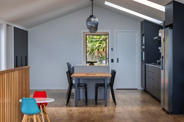 A contemporary open-plan kitchen and dining area with a compact timber dining table, dark dining chairs, and colourful children's chairs in the foreground. The room features dark full-height kitchen cabinetry, integrated appliances, a timber-slatted peninsula or screen, pale walls, a timber-look floor, a textured pendant light, and linear ceiling skylights or recessed lighting. Glazing and a door at the rear open toward leafy planting outside.