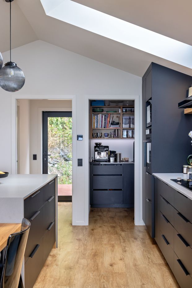 A contemporary kitchen with dark matte cabinetry, pale benchtops and warm timber flooring. The compact galley-style layout includes a central island with seating, a full-height bank of cabinetry, open shelving with a coffee machine and books, and a glazed exterior door at the rear. A large linear skylight brings abundant natural light into the space, complemented by a ribbed glass pendant.
