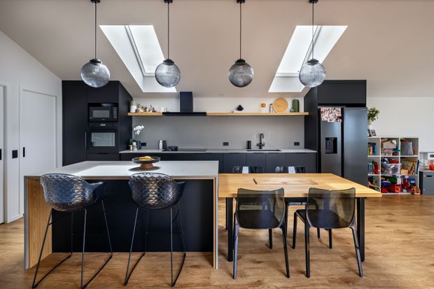 Bright contemporary kitchen and dining area with a central black island, integrated dark cabinetry, pale benchtops, timber dining table, transparent pendant lights, skylights, and warm timber flooring. A shelving and living area is visible beyond the kitchen.