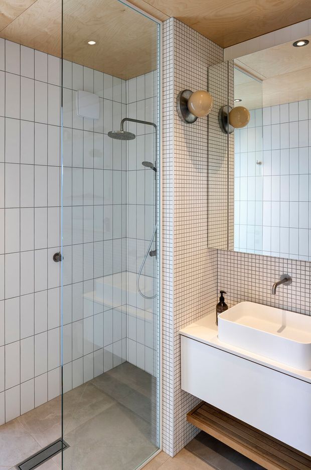 A contemporary bathroom with a frameless glass walk-in shower, wall-mounted rainfall and handheld shower fixtures, a white vessel basin on a floating vanity, and a large mirror. The walls are finished in large pale blue-grey rectangular tiles and a central vanity wall is covered in small square beige mosaic tiles. A warm timber plywood ceiling and timber shelf add contrast, while globe wall lights provide a refined mid-century-inspired detail.
