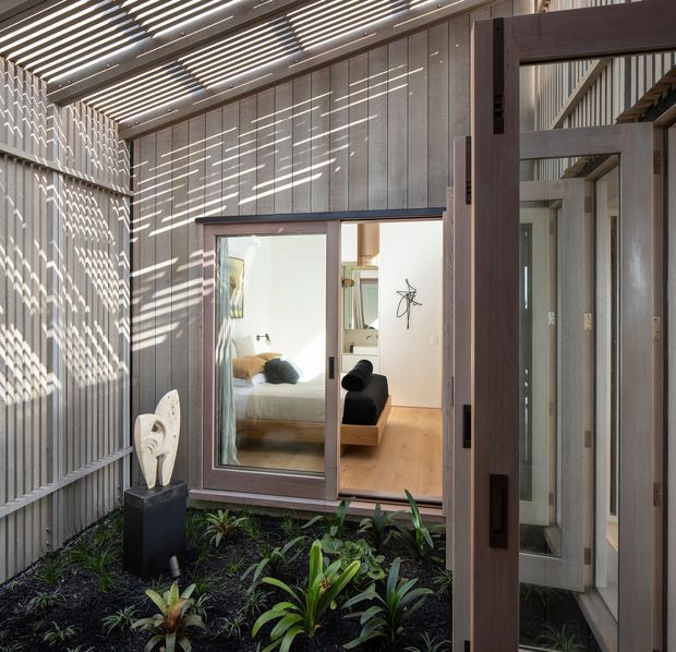 A sheltered internal courtyard beside a contemporary timber-clad dwelling, with a planted garden bed, sculptural feature, and large glazed sliding doors opening into a bedroom and adjacent interior. A slatted overhead canopy casts strong linear shadows across the walls and paving, while dark timber framing and vertical cladding create a warm, modern architectural character.