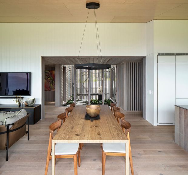 Bright contemporary open-plan dining area with a long natural timber dining table and coordinated wooden chairs, opening through a wide framed partition toward a planted outdoor courtyard. The room has pale timber flooring, white walls, a warm timber ceiling, vertical slatted screening, a dark linear pendant light, and built-in white cabinetry beside the kitchen. A living area with television and seating is visible to the left.