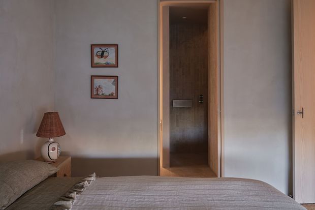 A softly lit bedroom with a neatly made bed in the foreground, warm neutral walls, a small timber bedside table with a fabric lampshade, and framed artwork. An open timber-framed doorway leads directly to a compact tiled shower room, while a timber door is visible at right. The overall scheme is understated, warm, and natural.