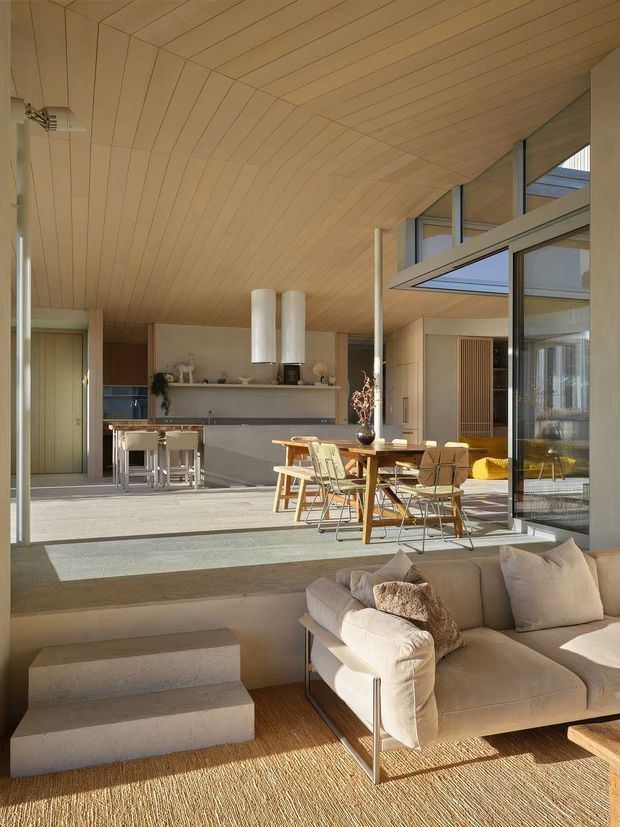 Sunlit open-plan interior with a contemporary kitchen, dining area and living room. The space features a warm light timber-lined ceiling, extensive glazing and clerestory windows, pale neutral finishes, a grey kitchen island, timber dining furniture, and a low-level stepped transition into the living area with a large cream sectional sofa.