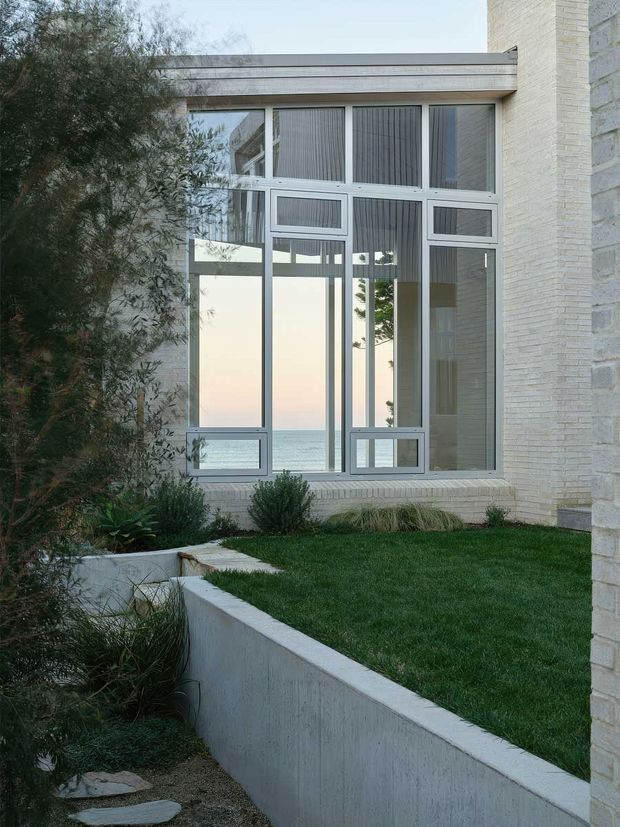 Exterior view of a contemporary light-brick building with a tall, multi-panel glazed opening and pale metal frames. The glazing reflects or reveals a coastal horizon, while a carefully landscaped courtyard foreground includes lawn, concrete retaining walls, gravel, stepping stones, and drought-tolerant planting.