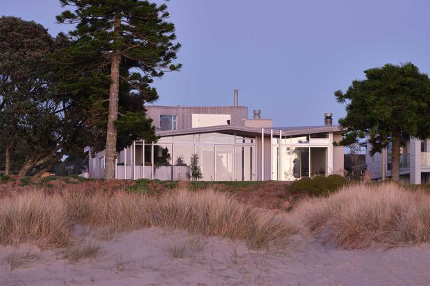 Exterior view of a low-rise contemporary coastal residence set behind natural sand dunes and grasses. The home has a pale, finely detailed facade, extensive glazing, slender vertical posts, a mostly flat roofline, and integrated outdoor-facing living areas. Mature wind-shaped trees frame the property, creating a calm beachfront architectural setting.