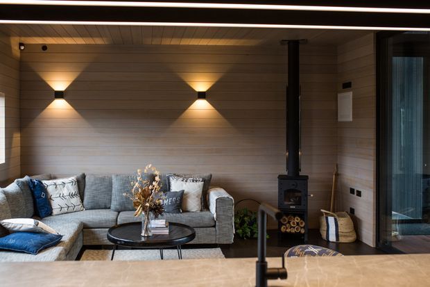 A warm, contemporary living room with a grey sectional sofa arranged around a round black coffee table. The room features horizontal timber wall and ceiling lining, integrated warm wall sconces, a black wood-burning stove with flue, and large glazed doors on the right. A timber-toned benchtop or island edge with a black tap is visible in the foreground, suggesting the living area adjoins an open-plan kitchen. The atmosphere is cozy and refined, with strong linear lighting and natural materials.