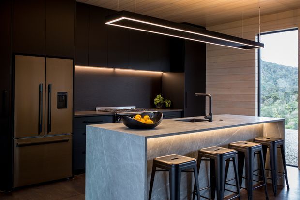 A sophisticated contemporary kitchen with a large stone-clad island and integrated seating for four. Dark, matte cabinetry and a matching refrigerator create a dramatic backdrop, while warm under-cabinet lighting and linear pendant lighting illuminate the island. The island features a dark sink and sculptural black tap, with timber wall panelling and expansive glazing on the right overlooking a green landscape.
