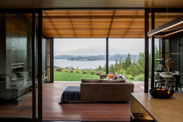 A contemporary open-plan living space with dark timber flooring and exposed timber ceiling slats, opening through large black-framed glazed doors to a landscaped lawn and expansive waterfront view. A brown sofa faces the opening, with a fireplace on the left and a dining area with linear pendant lighting on the right. The room has a warm, refined indoor-outdoor connection and strong horizontal framing.